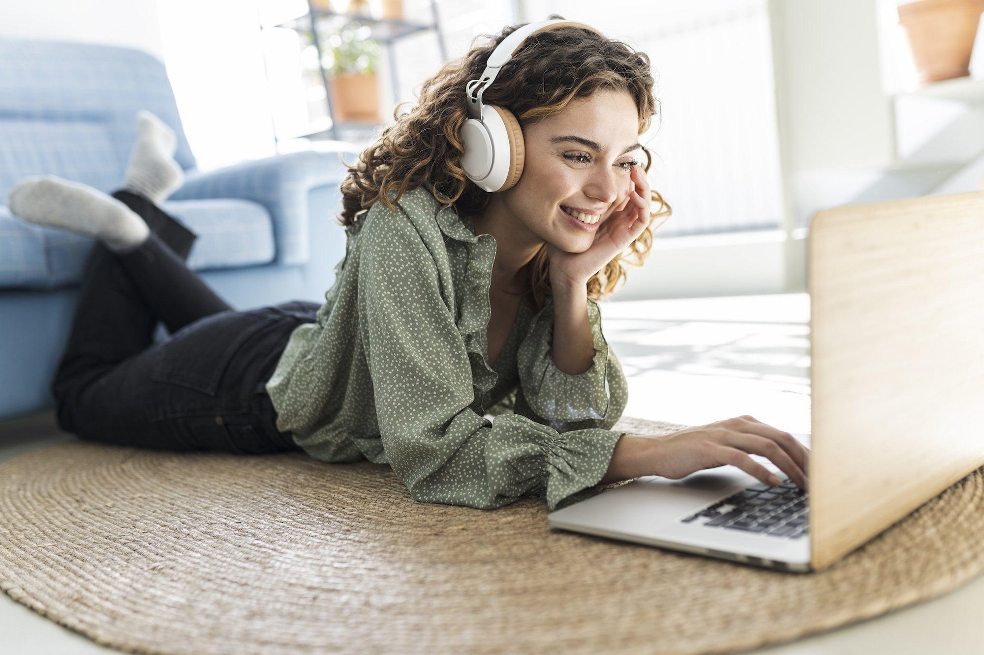 curly-haired woman lying on carpet at home using laptop, listening to music, studying.