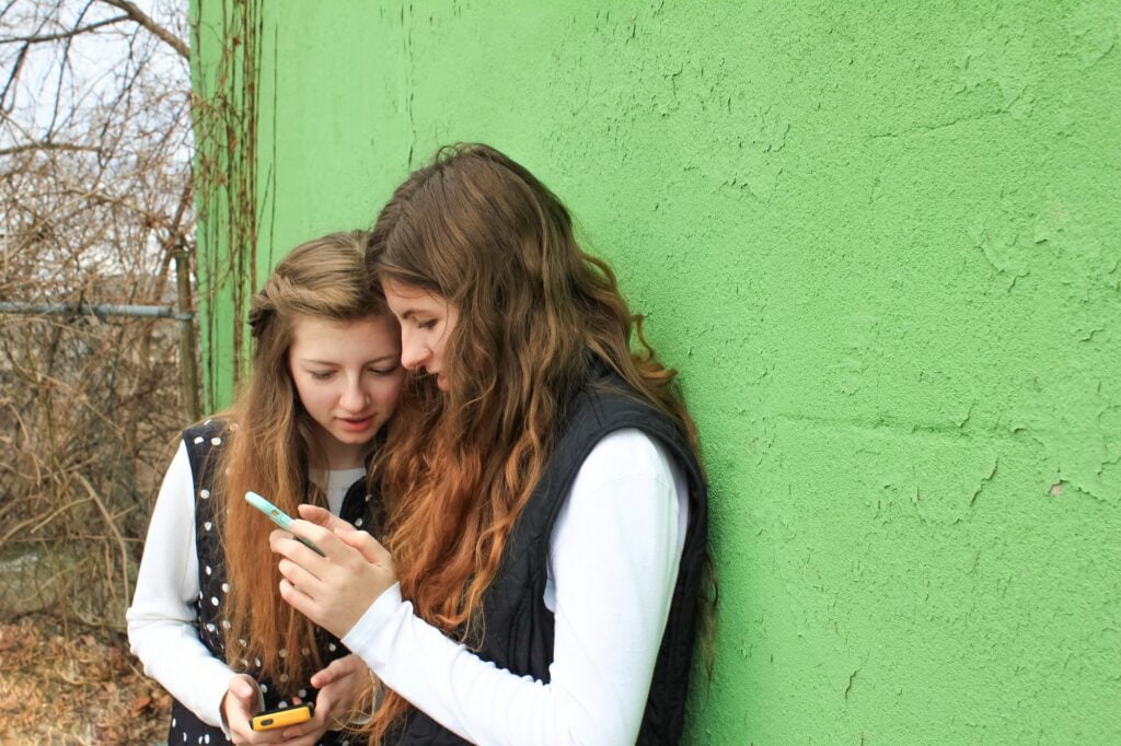 Two young women teens by green wall background using cell phones and reading social media posts